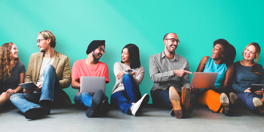 young adults of diverse cultures, backgrounds and ethnicities sit against aqua blue wall, talking and laughing