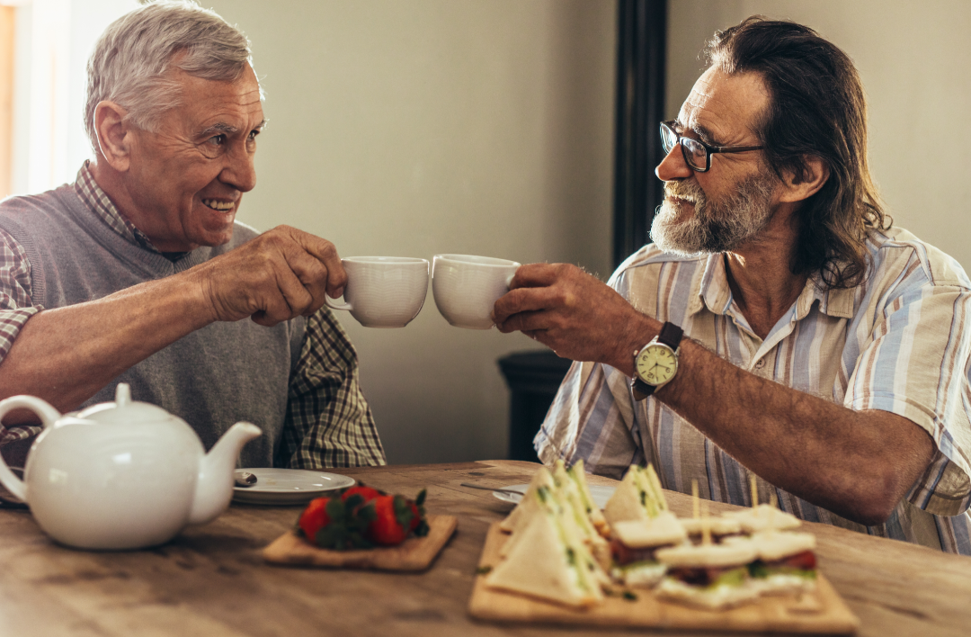 Elderly man in plaid and mature man with greying beard gesturing 'cheers' with tea cups